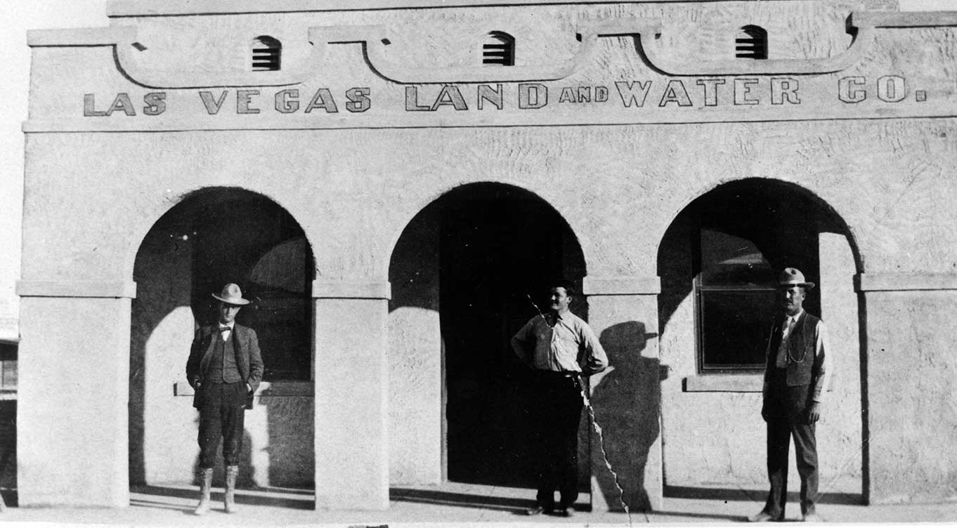 Three men standing in front of Las Vegas Land and Water Company building in grayscale photo.